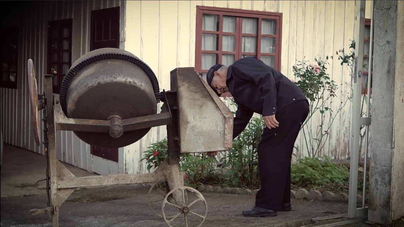 Senhor idoso observa betoneira antiga em frente a casa de madeira