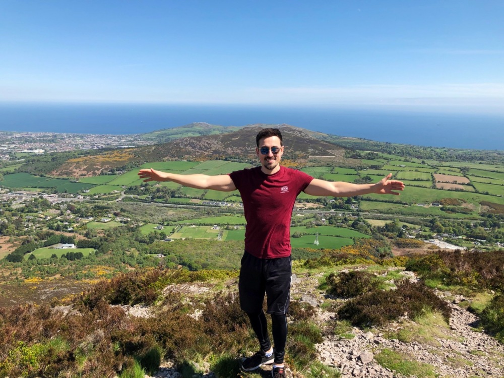 Marcelo com braços abertos em topo de montanha com vista panorâmica da costa da Irlanda