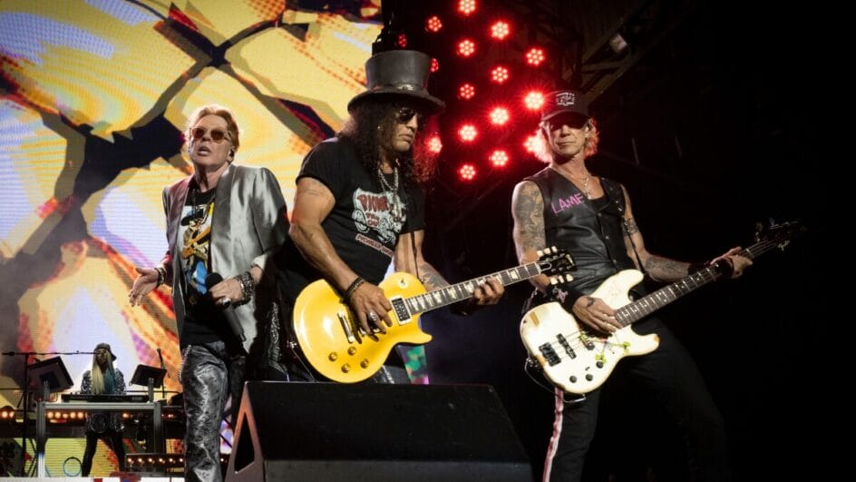 Axl Rose, Slash e Duff McKagan em apresentação ao vivo do Guns N’ Roses com luzes de palco ao fundo