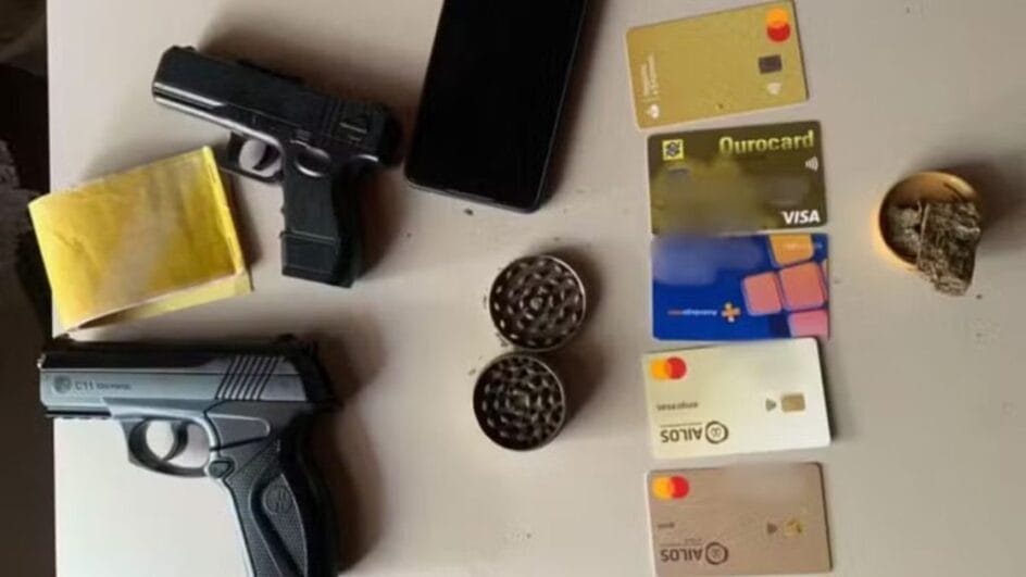 Objetos apreendidos incluindo duas armas de pressão, cartões bancários, celular e porção de maconha sobre uma mesa