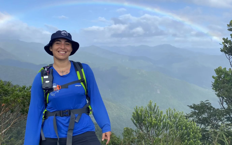Mulher sorridente com mochila e chapéu em uma trilha montanhosa, com arco-íris ao fundo, representando a jornada de Julieta Santamaria no Caminho da Mata Atlântica.