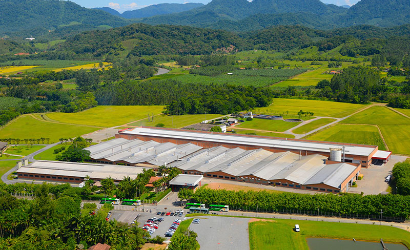 Vista aérea da unidade industrial em Corupá cercada por áreas verdes e montanhas ao fundo